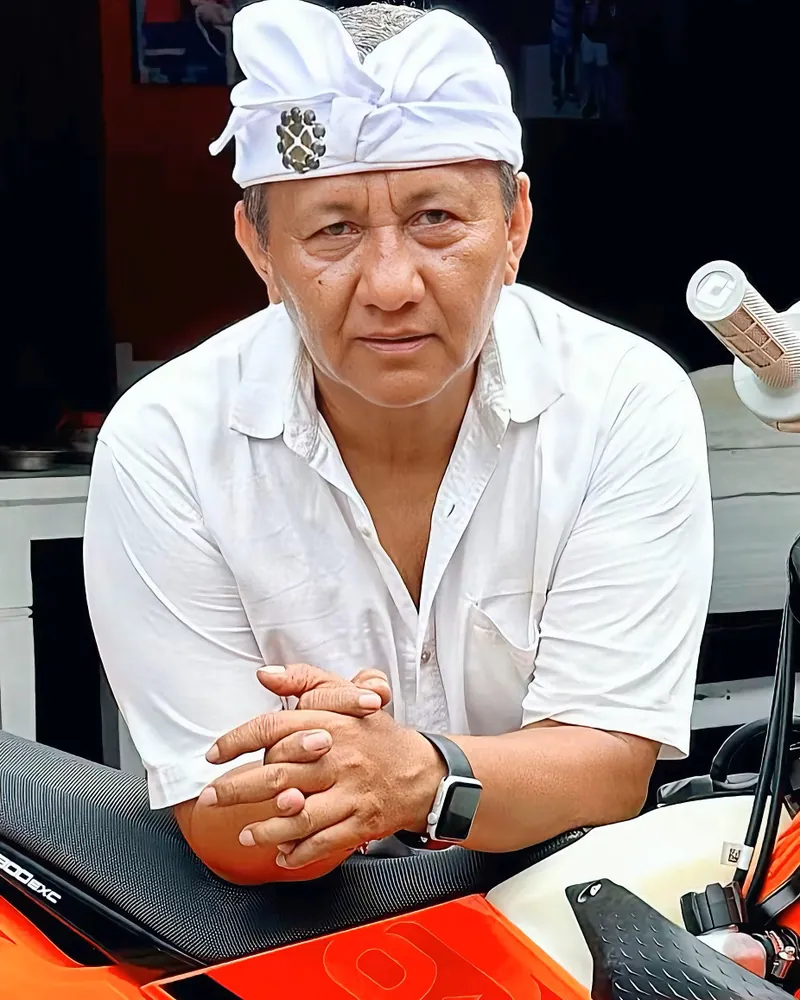 Portrait of Pak Putu, BaliEnduro's founder, in a traditional white Balinese udeng headdress and white linen shirt, resting his folded hands on the orange tank of a KTM enduro bike at the basecamp.